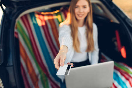 Travel and technology. Happy young woman working on laptop and holding credit card while sitting in trunk of carの写真素材
