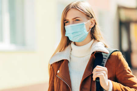 Beautiful stylish young woman wears a medical mask on a sunny city street, against the backdrop of a building,の写真素材