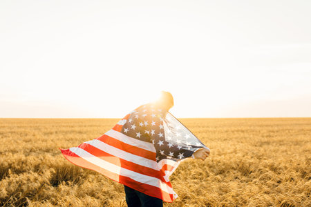 attractive man holding the flag of the United States against a blue sky on a clear sunny day, view from the back, close-up. National holiday conceptの写真素材