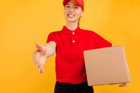 Delivery in red uniform holding a carton box parcel, Female employee working as a courier, on a yellow background, Service conceptの写真素材