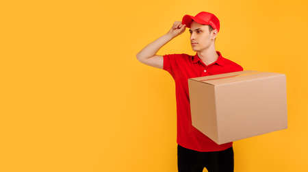 Happy excited male courier wearing red t-shirt and cap holding box parcel on isolated yellow...