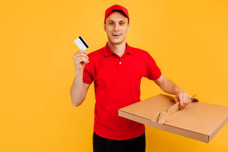 Delivery service man in red t-shirt and cap, gives food order and holds pizza box and credit card on...