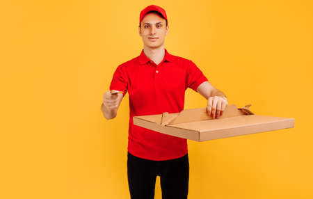 Delivery service man in red t-shirt and cap, gives food order and holds pizza box and credit card on isolated yellow backgroundの写真素材