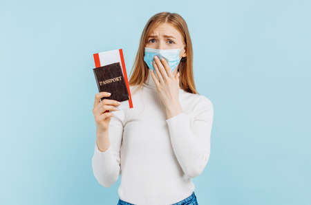 Shocked frightened young woman, in medical mask, holding passport with tickets, on blue backgroundの写真素材