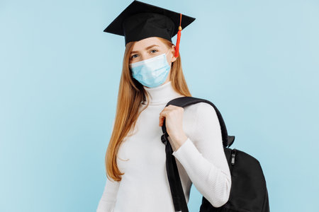 Happy young woman graduate in medical mask, student in graduation hat with briefcase on blue backgroundの写真素材