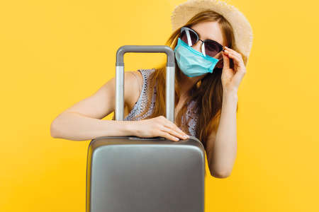 Young beautiful woman traveler, in a medical protective mask, in a summer dress and hat, with a suitcase on a yellow background, Passengers travel abroadの写真素材