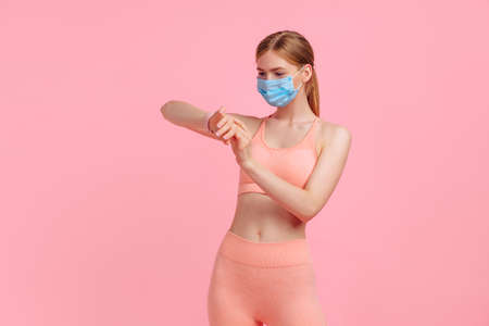 Young athletic woman wearing a protective mask against  virus disease, using a smartwatch, checking the activity of a wrist tracker, on a pink backgroundの写真素材