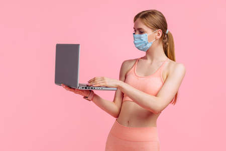Young sports fitness woman in a medical protective mask, in sportswear, uses a laptop on a pink background, healthy lifestyle conceptの写真素材