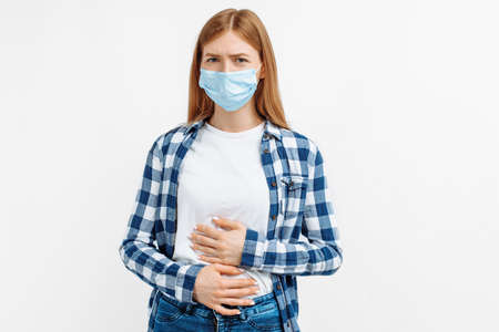 Young woman in protective mask holding both hands on her stomach while having stomach pain on isolated white backgroundの写真素材