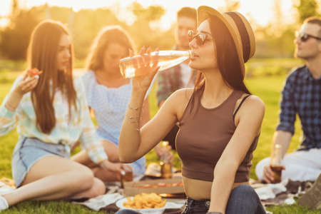 Beautiful young woman in a summer hat and glasses, resting with her friends, sitting on the lawn on a picnic in the park, summer picnicの写真素材