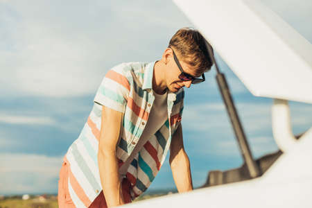 Breakdown of the car. Concentrated young man holding on to the hood of a car and looking inside while standing outdoors on a sunny dayの写真素材