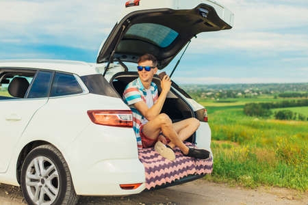 Happy young male traveler sitting in the trunk of his car, man having a rest during a trip, outdoors with a beautiful landscape, travel, active tourismの写真素材