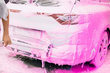 Car wash concept, Front view of white sports car covered with pink foam, Professional cleaning and car wash, Man washing car with high pressure waterの写真素材
