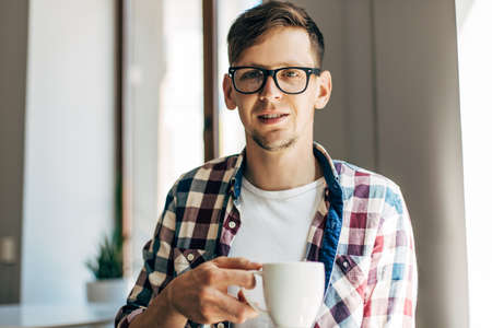 Happy young business man in glasses drinking aromatic coffee from a cup sitting in a cafe near the windowの写真素材