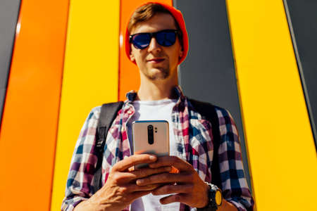Portrait of a handsome happy young man, wearing sunglasses and a hat, using his mobile phone,...