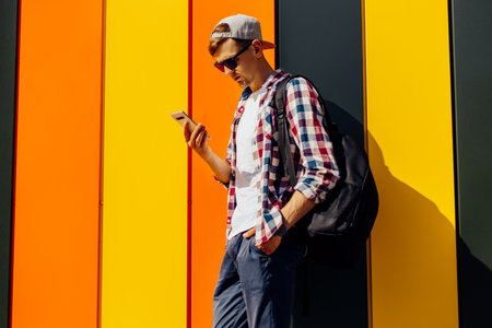 Young smiling stylish man in sunglasses and a cap, uses a mobile phone, leaning against a colorful...