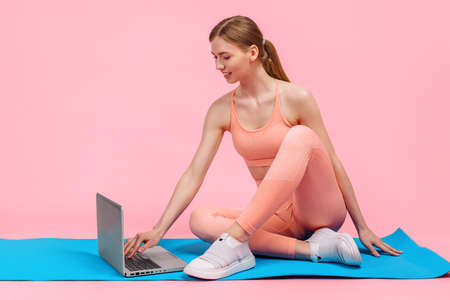 Fitness training online, young sportive woman doing workout with laptop, on pink backgroundの写真素材