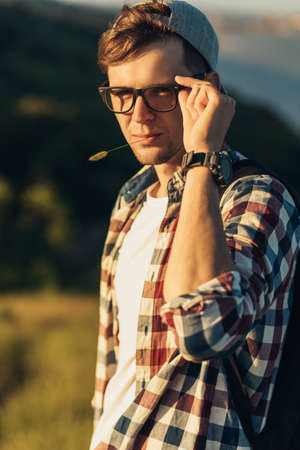 Portrait of a young handsome man, wearing glasses and a cap, smiling young man, standing on the street in nature, against the background of a beautiful forestの写真素材