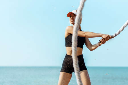 Beautiful young sportive woman, the athlete moves the ropes in wave motion as part of the training for burning fat, training on the city beach, Sportswoman training with fighting ropes by the seaの写真素材