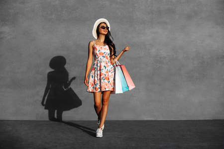 Stylish woman, wearing a hat and sunglasses, walks through the city streets with shopping bags,...