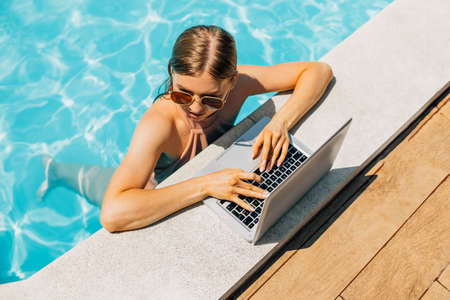 Attractive young woman in sunglasses, uses laptop and work remotely over the pool, Attractive woman smiling, using laptop in the pool, Summer vacation concept, remote workの写真素材