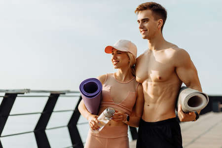 Young fitness couple go to workout, man and woman with fitness mats in hands, getting ready to workout outdoors on the pier, sport and healthy lifestyleの写真素材