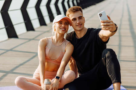Attractive young sportsmen man and woman resting after workout taking selfie on mobile phone sitting on fitness mat outdoors on pierの写真素材