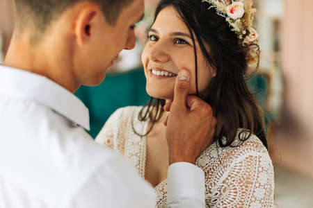 Happy young couple, stylish groom in a suit and a young bride in a wedding lace dress with a bouquet of flowers, sit together, hug, in the studio on the sofa, wedding in boho styleの写真素材