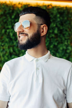 Portrait of a happy young man with a beard, in a white shirt and sunglasses, against a background of...