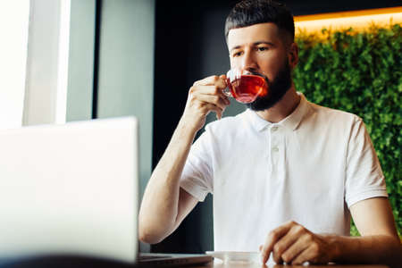 Smiling young bearded man in glasses using a laptop in a cafe and drinking tea while sitting at a wooden table, Male hands are typing on the laptop keyboard, Concept of young people working mobile devicesの写真素材