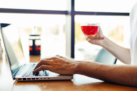 man using a laptop in a cafe and drinking tea while sitting at a wooden table, Male hands are typing on the laptop keyboardの写真素材