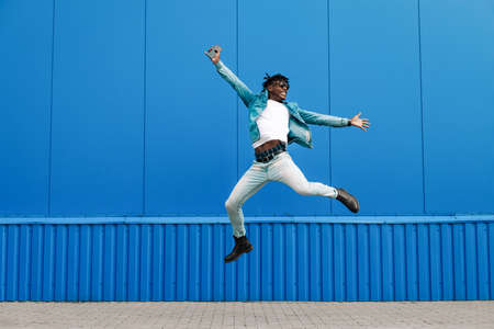 african guy jumping with a phone in his hands, in the city against the background of a blue building, concept of successの写真素材