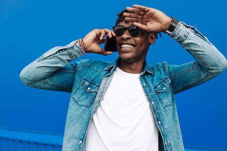 african man in sunglasses typing sms over blue background, 5g internet concept, showing gesture of tiredness, black man in blue shirt laughing with a phone in his handsの写真素材