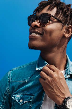 fashion african man in sunglasses in trendy modern clothes posing against the background of a blue...