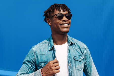 fashion african man in sunglasses in trendy modern clothes posing against the background of a blue building, on the streetの写真素材