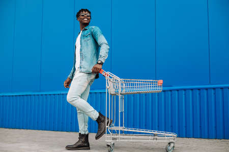 black afro guy shopping with a shopping cart, guy posing, fashion fashion, shopping near the mall, young black student on the background of a blue buildingの写真素材