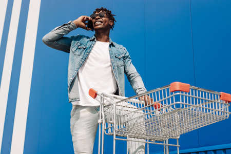 african black man walking with a shopping cart talking on the phone, making a phone call, shopping online talking with an operator, against background of a blue buildingの写真素材