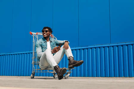 Afro black man with a shopping cart and a phone in his hands, makes purchases online, against background of a blue buildingの写真素材