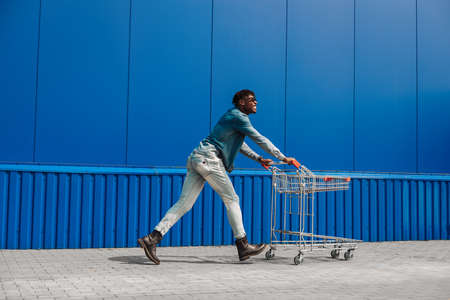 black afro guy runs to shop with a shopping cart, the guy has fun and jumps, runs for purchases near the mall, young black student on background of a blue buildingの写真素材