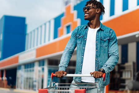 afro american black man with shopping trolley in front of the mall, shopping in the mall, successful shopping mall, wearing goggles in sunの写真素材