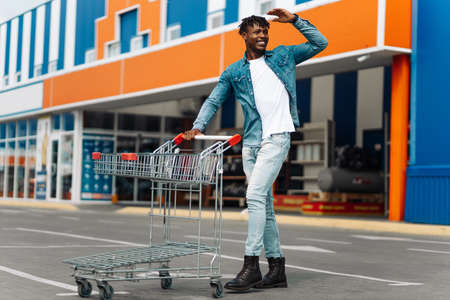 afro american black man with shopping cart in front of mall shopping mall, successful shopping mall, looks into the distanceの写真素材