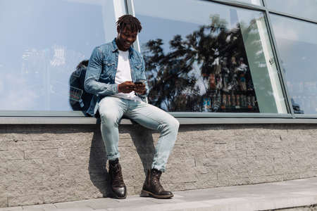 black man African American with a phone in his hands, typing an SMS message, sitting against an educational institution, in the city center, against a glass building, cheerful Africanの写真素材