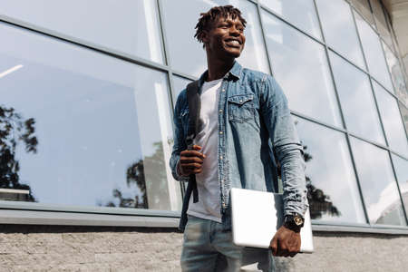 a dark-skinned African American with a laptop in his hands walks down the street against a stele...
