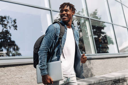 a dark-skinned African American with a laptop in his hands walks down the street against a stele building, in the city centerの写真素材