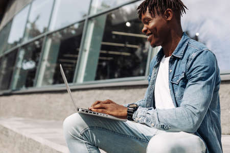 black African American typing on a laptop on the street in front of a glass building, sitting online, working on the road, high-speed mobile internet, 5g, 4gの写真素材