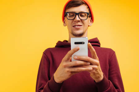 A handsome young guy in an orange hat with glasses and a red sweater is typing a message on the phone, holding the phone in his hands, standing on a yellow background, autumnal concept, 5gの写真素材