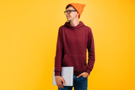 young handsome guy with a laptop in his hands, in an orange hat with glasses and a red sweater, on a yellow background, autumn conceptの写真素材
