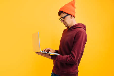 young handsome guy with typing on a laptop, in an orange hat with glasses and a red sweater, on a yellow background, high-speed internet, 5gの写真素材