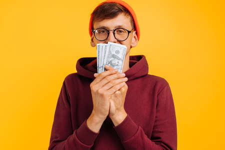 handsome man wearing an orange hat with glasses and a red sweatshirt, sniffing a dollar bill,...