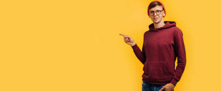 man on yellow background points to blank space, in orange cap and red sweater, isolated for text,...